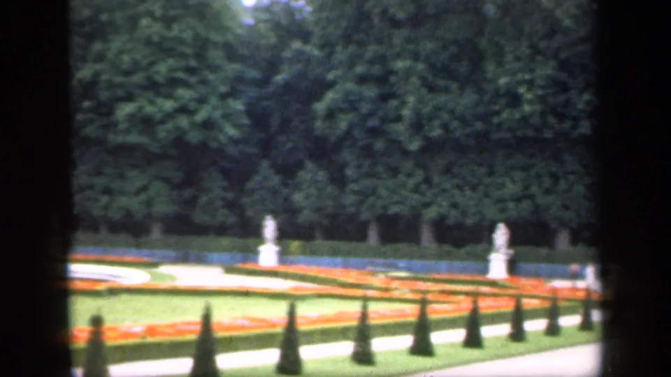 1949 Ireland 8mm view across a formal garden with orange beds, trimmed cone shrubs, pale paths, statues, lawns, and mature trees.