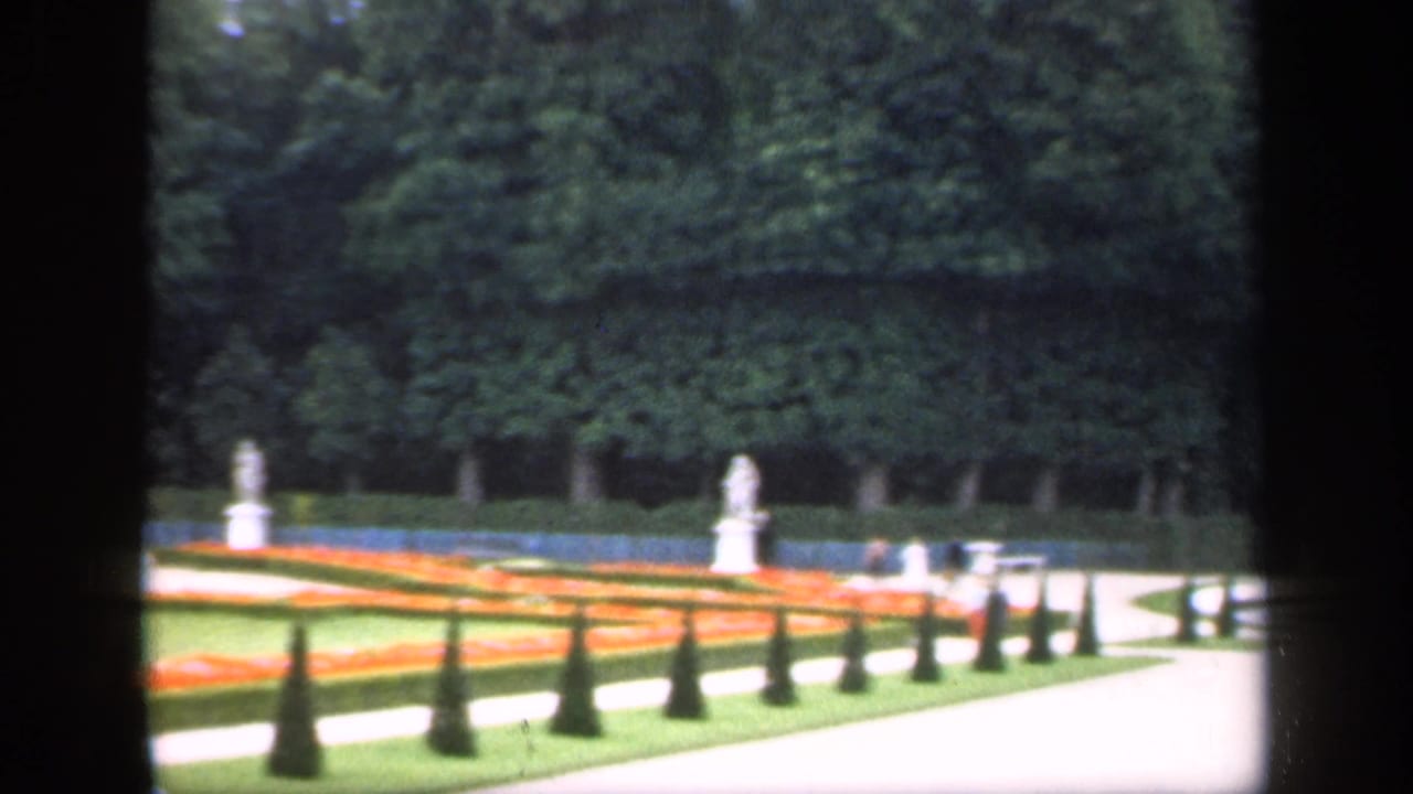 Formal garden with red flowerbeds, pale paths, statues, trees, and dark vertical framing.