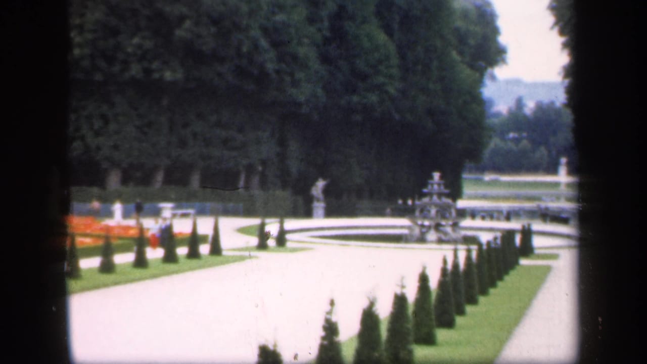 Curving garden path with conical hedges, fountains, lawns, trees, and distant visitors.