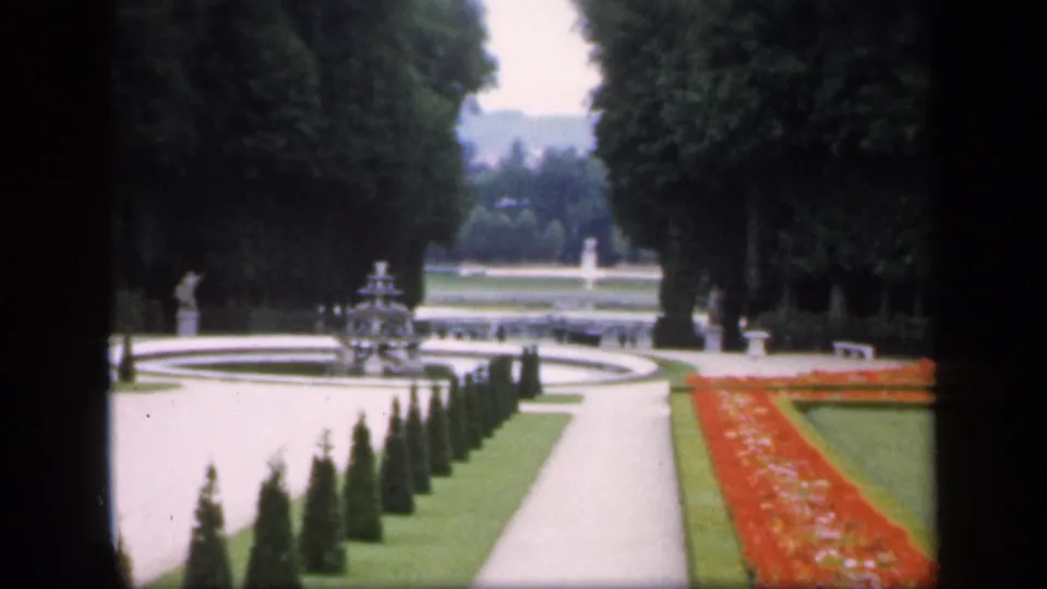 1949 Ireland 8mm frame with clipped hedges, a pale path, fountain area, red flowerbed, trees, and small distant figures.