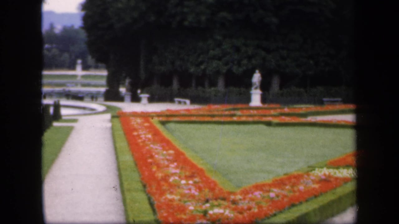 Formal garden with red flowerbeds, green lawn, pale paths, clipped hedges, trees, and stone statues.