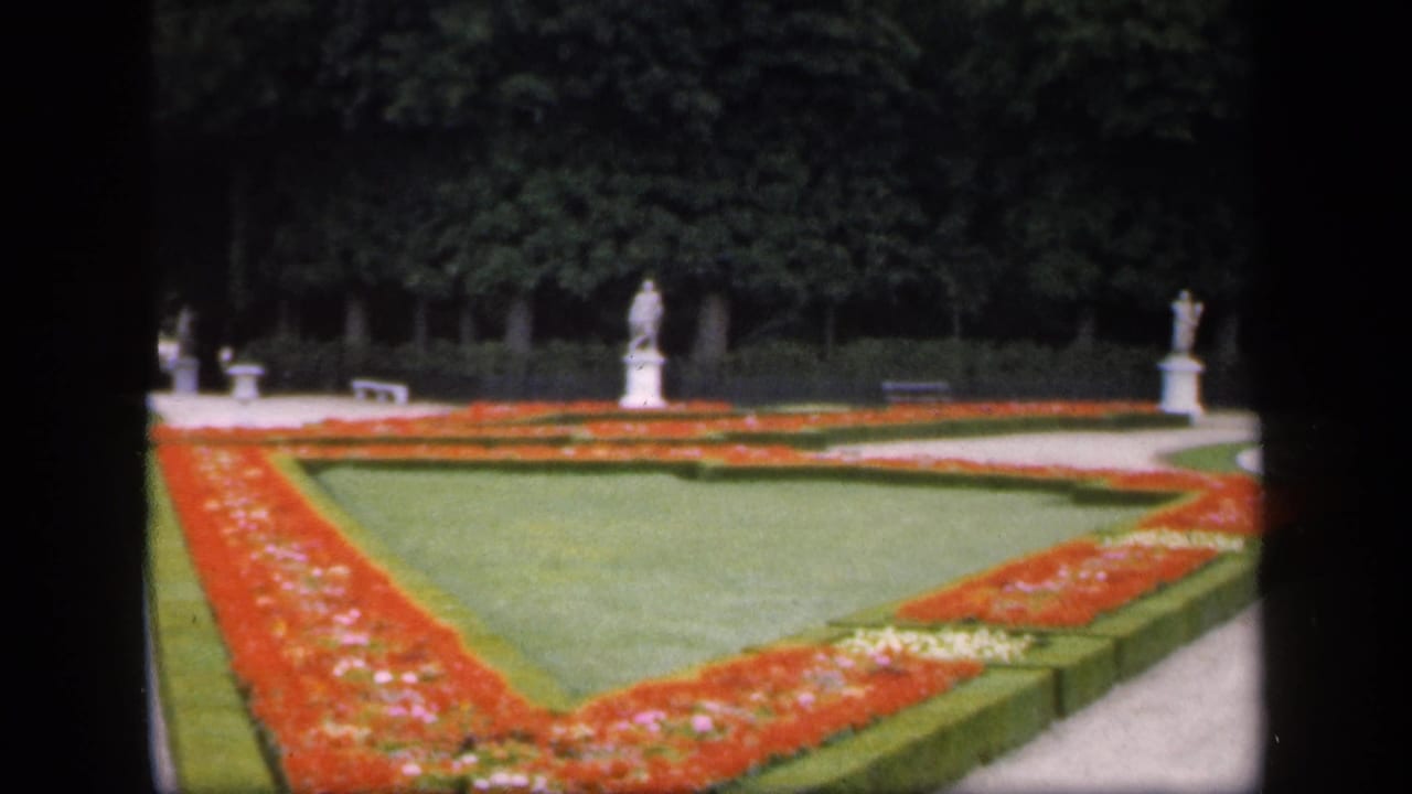 Geometric red flowerbeds and lawns bordered by hedges, with trees, benches, and stone statues in the background.