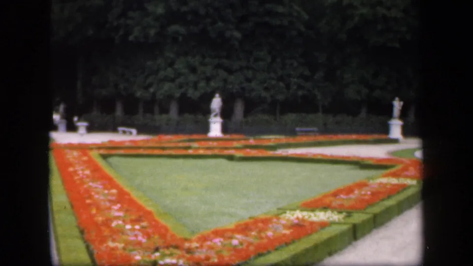 1949 Ireland garden view with red and white flowerbeds arranged around lawns, backed by clipped hedges, trees, benches, and pale stone statues.