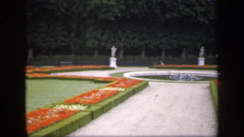 1949 Ireland 8mm frame showing a pale garden path, red flowerbeds, clipped hedges, green lawns, tree line, and a circular fountain basin.