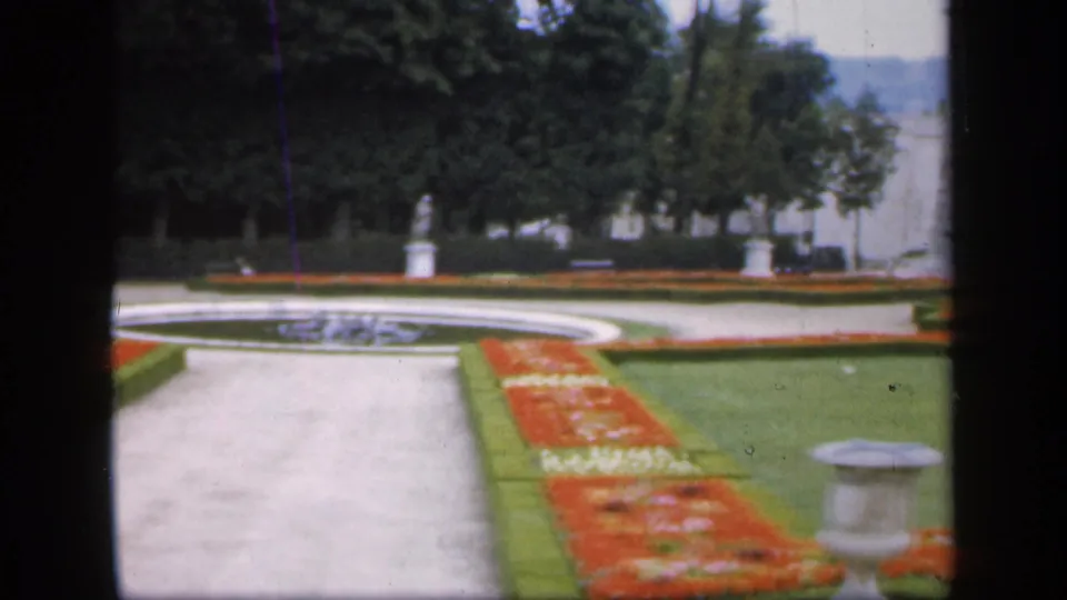 1949 Ireland garden frame with a circular fountain, pale path, red flowerbeds, green lawns, distant statues, trees, and a stone urn or pedestal.
