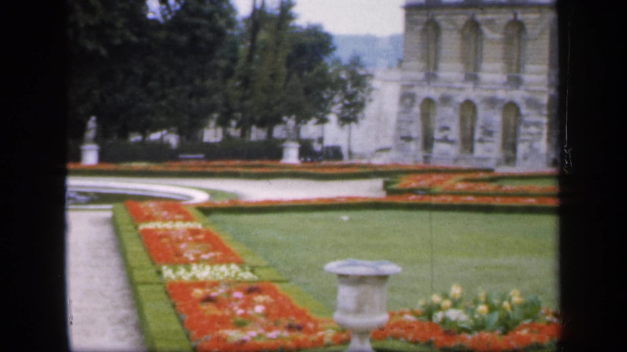 Formal garden with flowerbeds, lawn, urn, trees, and pale stone building beyond.