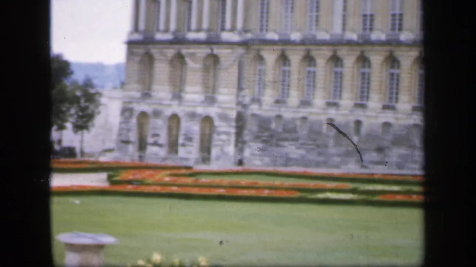 1949 Ireland home-movie frame with geometric red flowerbeds, open lawn, trees, and a large classical stone building in the background.