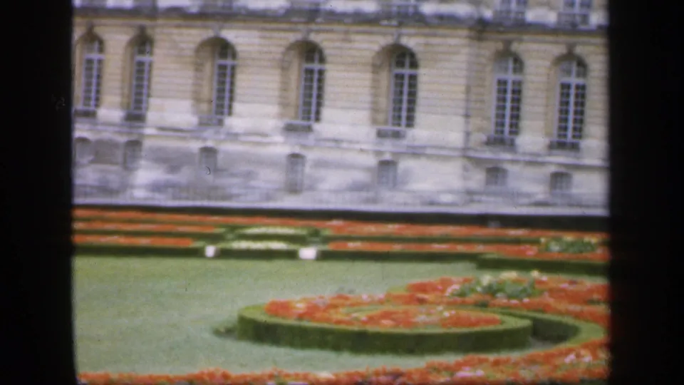 1949 Ireland frame with a curved formal flowerbed in the foreground, manicured lawn, and a pale historic building facade behind it.