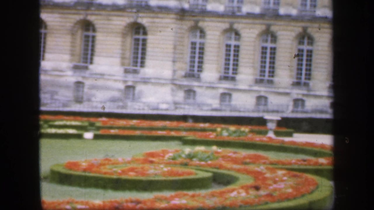 Formal flowerbeds and clipped hedges in front of a pale stone building with tall arched windows.