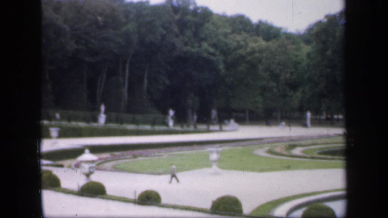 Formal garden paths, lawns, white statuary, trees, and a distant person walking.