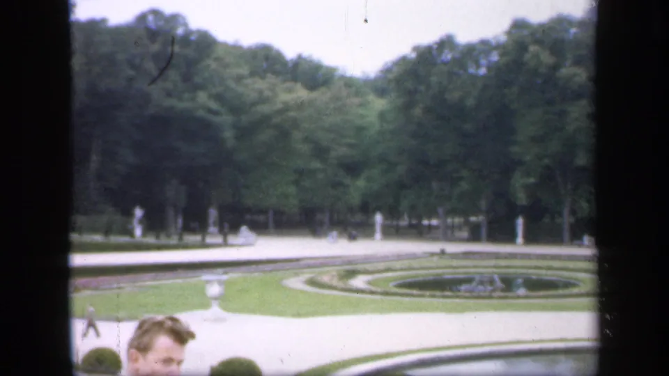 1949 Ireland 8mm frame showing curved formal beds, gravel paths, trees, white statuary, and a partly visible visitor near the camera.