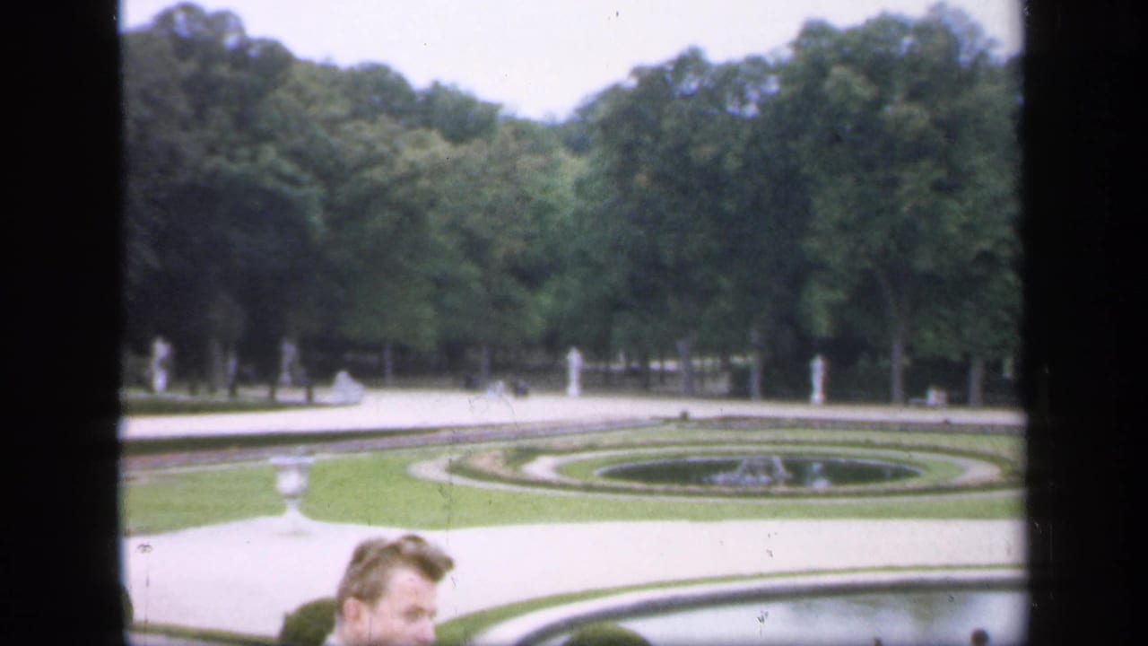 1949 Ireland 8mm frame showing a formal garden with circular lawns, a fountain basin, dense trees, paths, and visitors partly visible at the bottom.