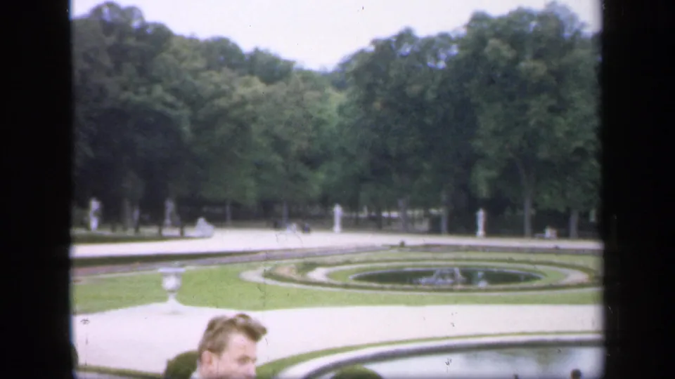 1949 Ireland 8mm frame showing a formal garden with circular lawns, a fountain basin, dense trees, paths, and visitors partly visible at the bottom.