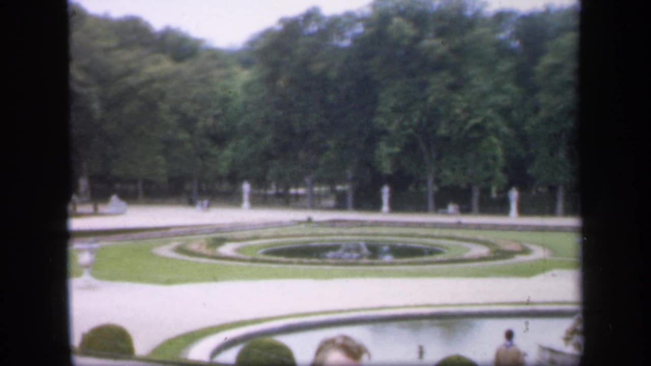 Formal garden seen across paths, circular lawn beds, trees, statues, and partial visitors.