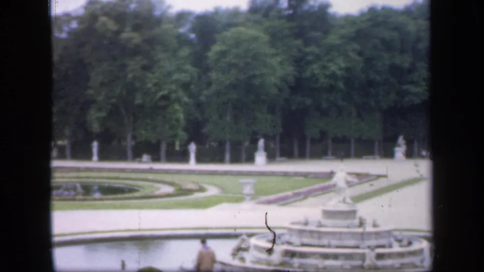 1949 Ireland 8mm frame showing foreground water, a tiered fountain at right, circular garden beds, statues, dense trees, and a visitor seen from behind.
