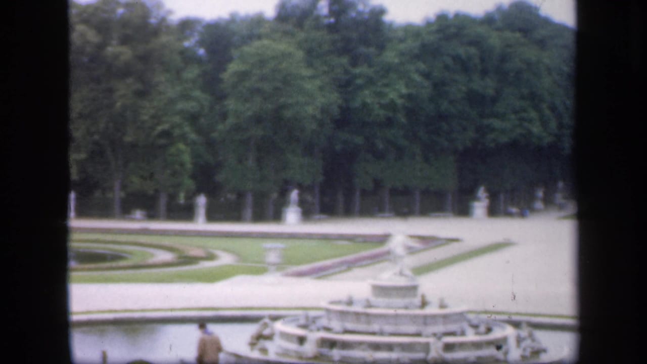 Elevated view of an ornate fountain, pale paths, clipped lawns, statues, and dense trees.