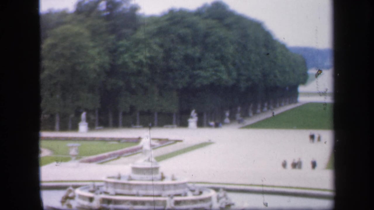 Fountain garden with symmetrical paths, dense trees, statues, lawns, and small distant visitors.