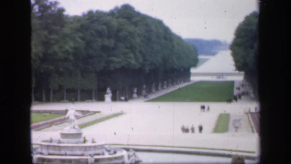 Elevated 1949 8mm view in Ireland showing a tree-lined avenue, fountain edge, pale paths, lawns, statues, and groups of visitors.