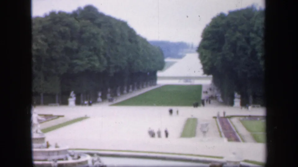 1949 Ireland 8mm frame of a formal garden avenue with clipped trees, lawns, statues, flower beds, pale paths, and distant visitors.