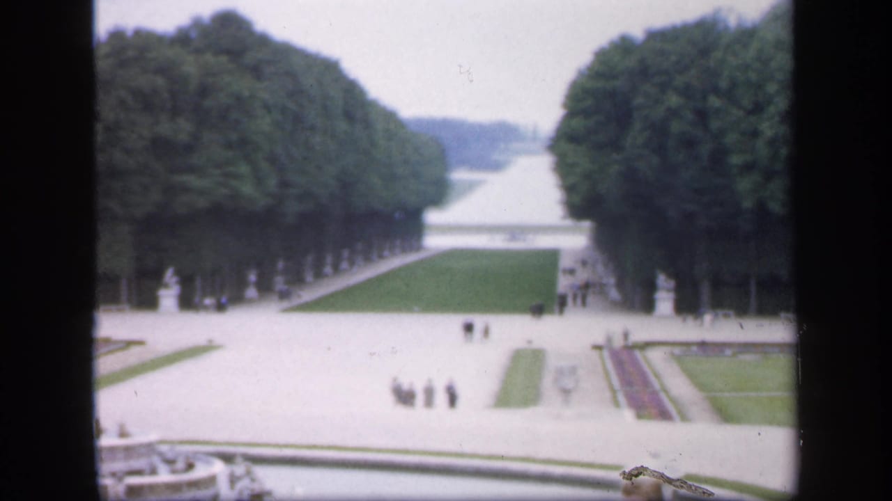 Formal garden seen through dark window framing, with tree-lined paths, lawns, statues, and distant visitors.