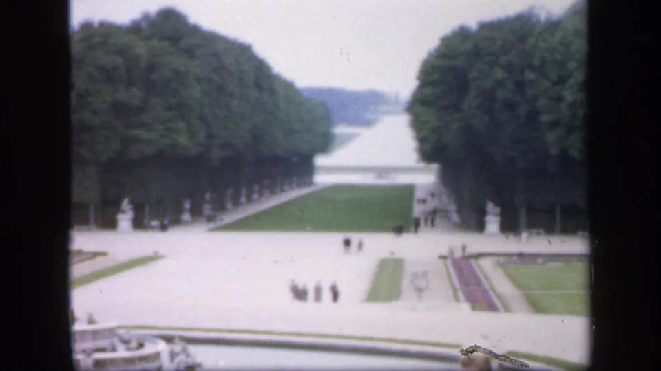 1949 Ireland 8mm view of a formal garden from behind a dark window frame, showing clipped lawns, avenues, statues, and small groups on paths.