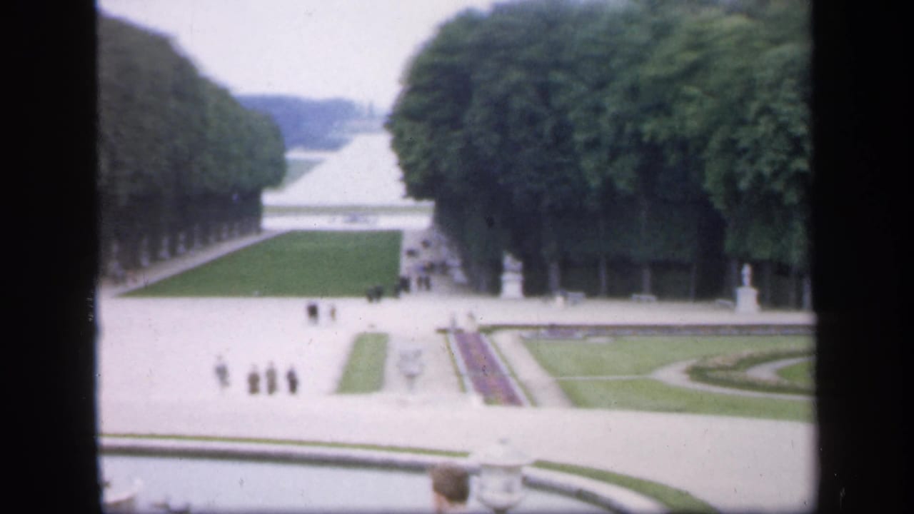 Soft 8mm frame of a formal Irish garden in 1949, with broad paths, statues, clipped lawns, flowerbeds, and distant pedestrians.