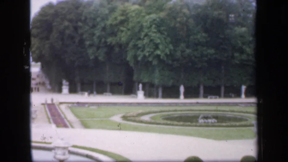 Soft 8mm view of a formal garden in Ireland, 1949, with circular lawn design, statues, broad paths, dense trees, and small figures.
