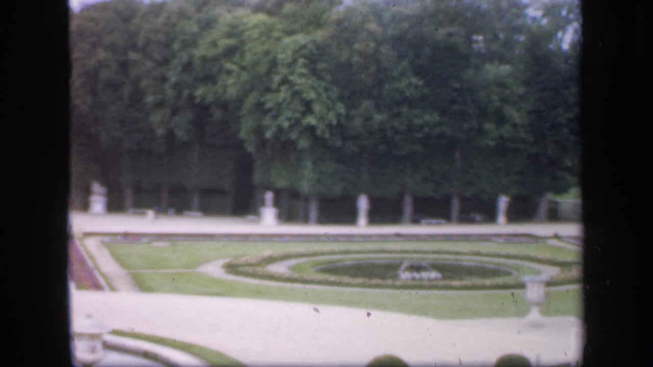 1949 Ireland home-movie frame showing a formal garden with circular lawn beds, pale paths, white statues, urns, and dense trees.