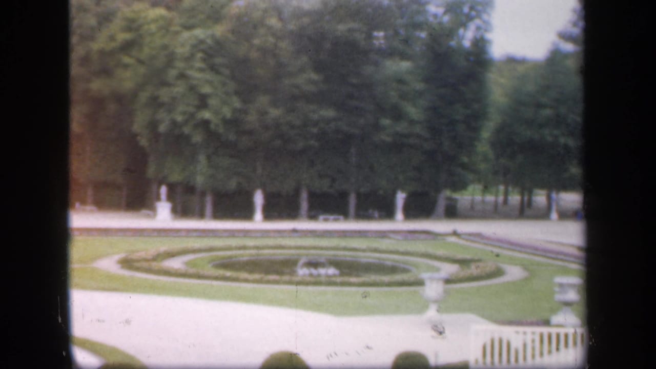 Formal garden pan with circular beds, pale walkway, statues, urn, trees, and low fence.