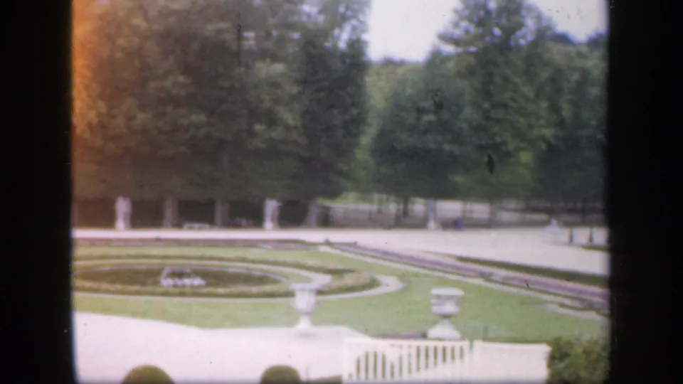 1949 Ireland garden view with circular clipped lawn, urn planters, white statues, tree backdrop, and visible film wear along the frame edge.