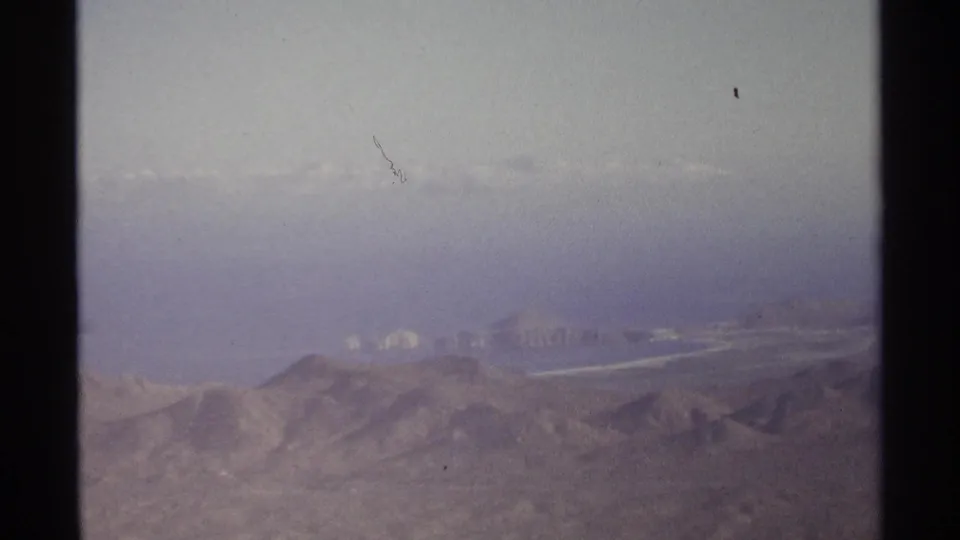 1974 home-movie frame near Cabo San Lucas showing arid hills and faint coastal forms beyond the haze.