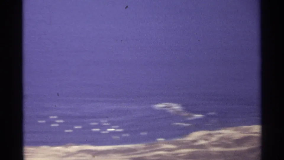 1974 Cabo San Lucas aerial frame showing blue ocean, pale coastal edge, and scattered white surf or buoy-like marks.