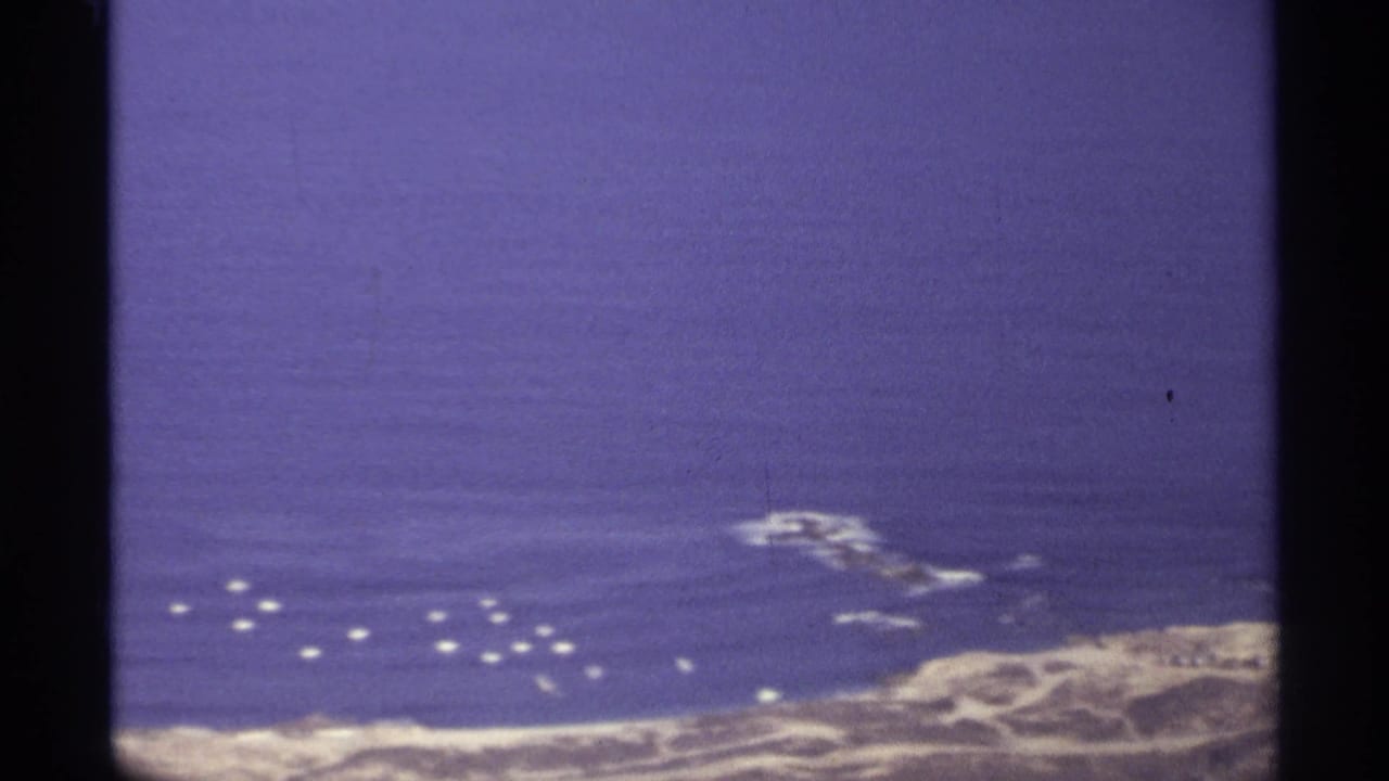 Aerial view of blue sea, rocky shoreline, and small white objects offshore.