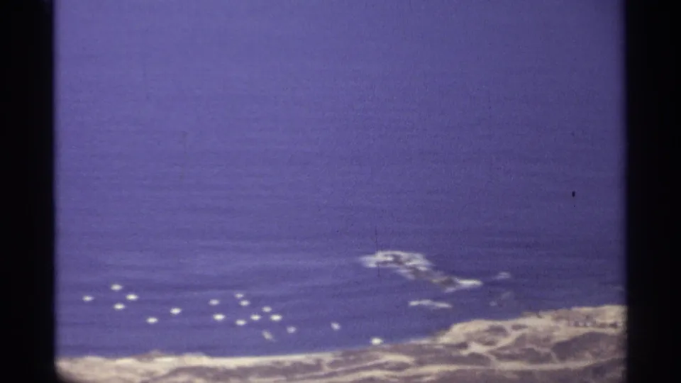 1974 Cabo San Lucas 8mm aerial view showing blue water, pale surf, rocky coast, and small white buoys or boats offshore.