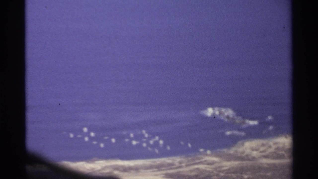 High-angle sea view with rocky coast and a line of small white offshore objects.