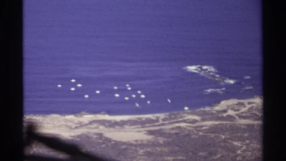 1974 Cabo San Lucas aerial home-movie frame with blue sea, rocky shoreline, pale surf, and scattered white buoys or boats.