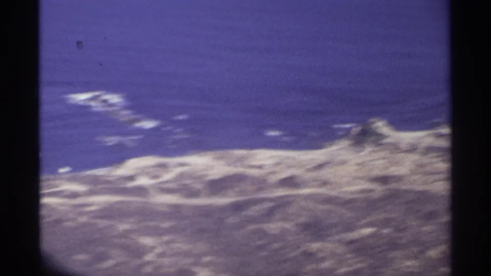 1974 aerial home-movie frame from Cabo San Lucas with rugged coast, blue water, and dark airplane-window dividers.
