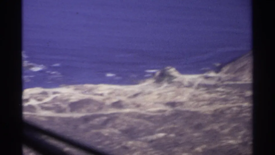 Aerial 8mm view of Cabo San Lucas coastline in 1974, with pale landforms, surf lines, and ocean beyond the window.