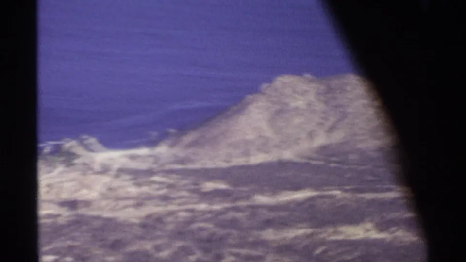 1974 Cabo San Lucas plane-window view with blue ocean, rocky desert coast, and dark frame edge from the aircraft interior.