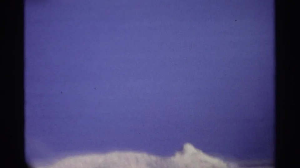 1974 8mm frame with a distant snow-covered peak near the lower edge and broad blue sky above.