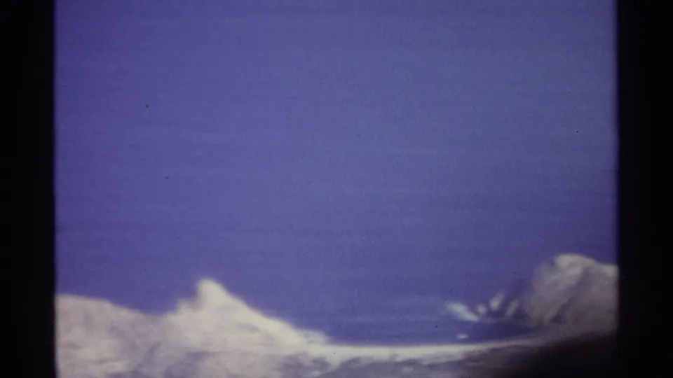 1974 8mm frame with a snow-covered slope and peak at lower right, framed by blue sky and film-edge shadow.