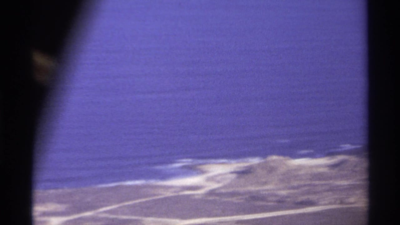 Aerial view of blue ocean and pale coastline seen through a dark airplane window frame.