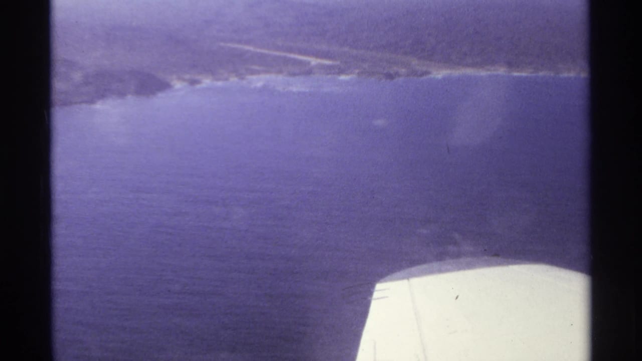 1974 Cabo San Lucas aerial frame with blue ocean, distant shore, and the white airplane wing visible at lower right.
