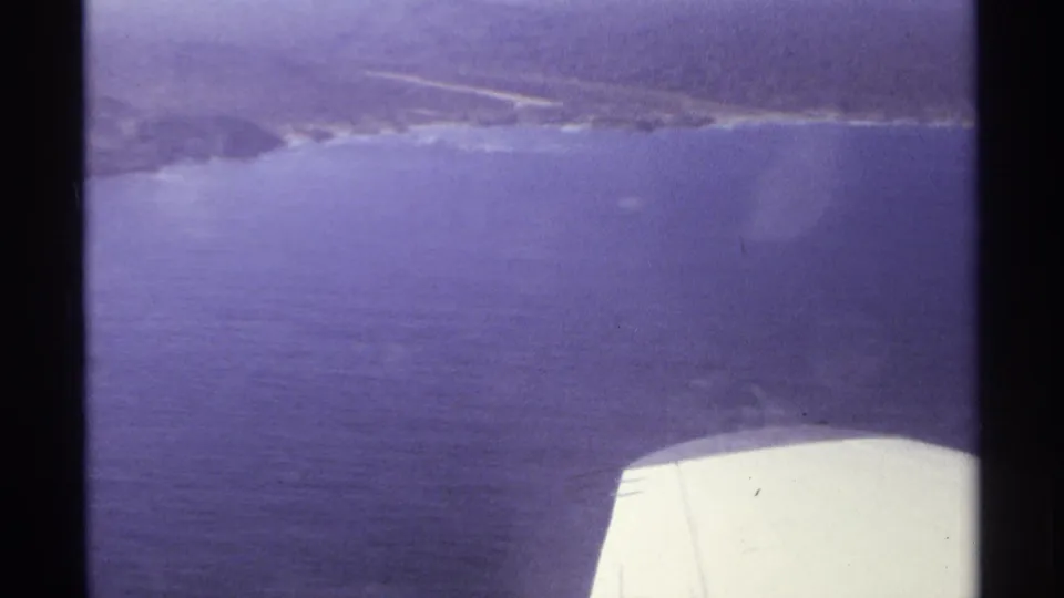 1974 Cabo San Lucas aerial frame with blue ocean, distant shore, and the white airplane wing visible at lower right.