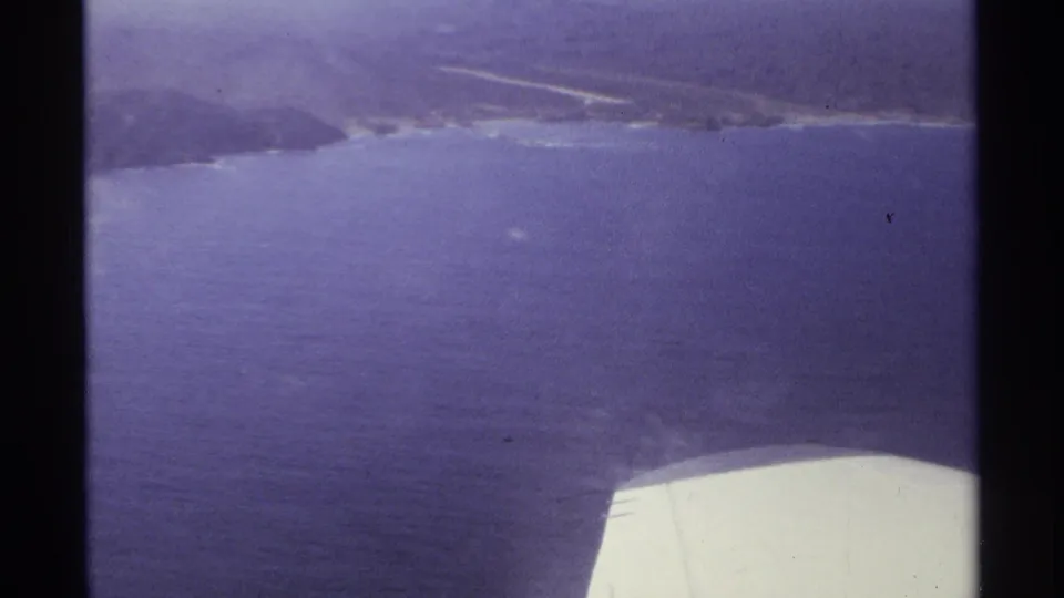 1974 Cabo San Lucas frame showing the aircraft wing below a blue coastal expanse and distant shoreline.