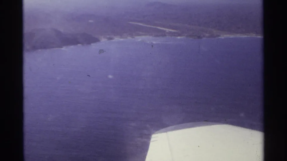 1974 Cabo San Lucas aerial frame with hazy coastline, blue ocean, and the airplane wing along the lower edge.