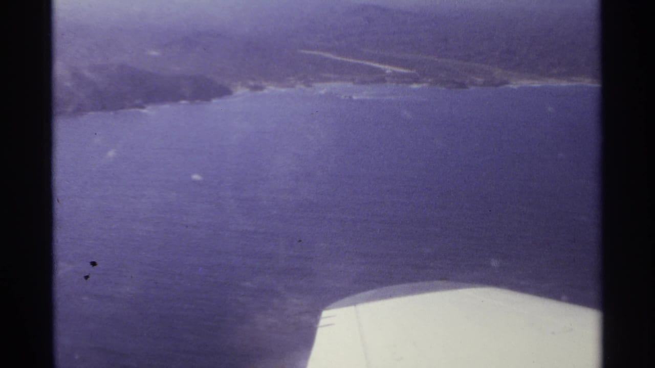 Aerial view over blue water and coastline with an airplane wing at the bottom edge.