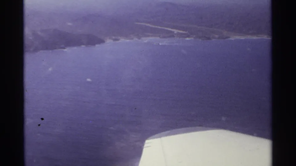 1974 8mm view from an airplane over Cabo San Lucas coastline, with ocean, shoreline, hills, and a wing visible.