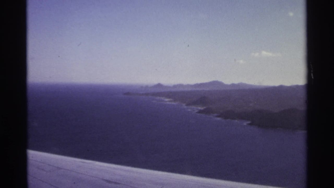 Airplane wing in the foreground with ocean, coastline, and low mountains beyond.