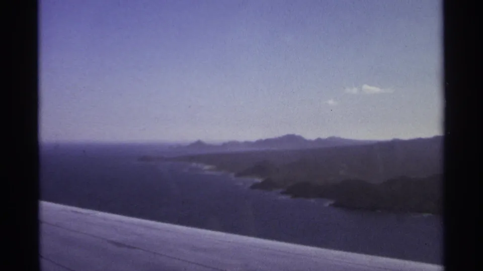 1974 home-movie aerial view of Cabo San Lucas coast, with wing, sea, peninsula, and hazy mountains.